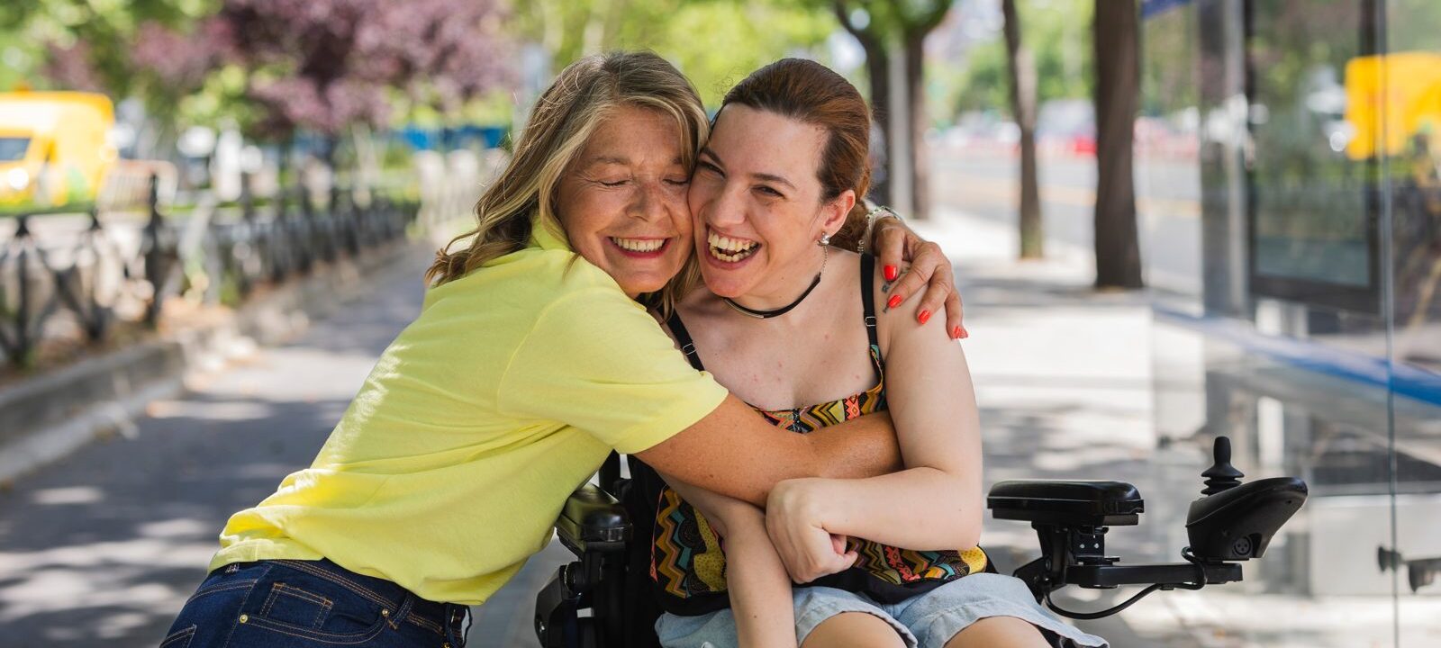 Smiling woman in a yellow top hugging a friend who uses a power wheelchair on a tree-lined street
