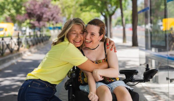 Smiling woman in a yellow top hugging a friend who uses a power wheelchair on a tree-lined street