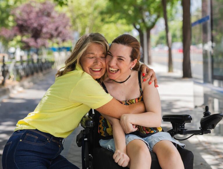 Smiling woman in a yellow top hugging a friend who uses a power wheelchair on a tree-lined street